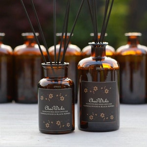 May include: Two brown glass bottles with black labels and gold lettering. The labels read "Chad Wick's Hand Poured Reed Diffuser - Sandalwood & Black Pepper". The bottles are filled with a clear liquid and have black reed sticks sticking out of the top.