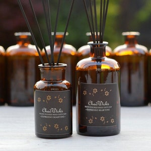 May include: Two brown glass bottles with black reed diffusers and labels that read "Chad Wick's Hand Poured Reed Diffuser - Espresso Martini - ". The bottles are sitting on a wooden surface.