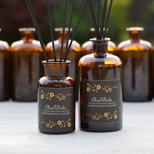 May include: Two brown glass bottles with black labels and gold accents. The labels read "Chad Wick's Hand Poured Reed Diffuser - Lemongrass & Ginger - ". The bottles are filled with a clear liquid and have black reed sticks sticking out of them.