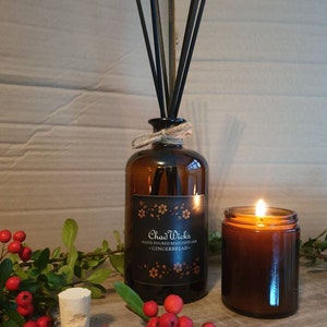 May include: A brown glass reed diffuser with black reeds and a matching candle. The diffuser has a label that reads "Chad Wicks Hand Poured Reed Diffuser - Gingerbread." Red berries and greenery are arranged around the products.