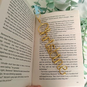 May include: A gold-coloured wire bookmark shaped into the cursive word "Imagine" with a light blue ribbon attached to the top loop. The bookmark is placed vertically on an open book page with printed black text, showing a portion of a story.