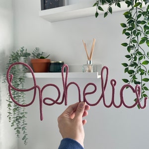 以下が含まれることがあります： 白い壁に掛けられた、ピンクのロープで作った「オフィーリア」という文字のウォールアートです。