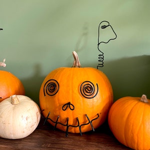 May include: A carved orange pumpkin with a wire ghost and a wire face on a wooden shelf. There are two other pumpkins on the shelf, one white and one orange.