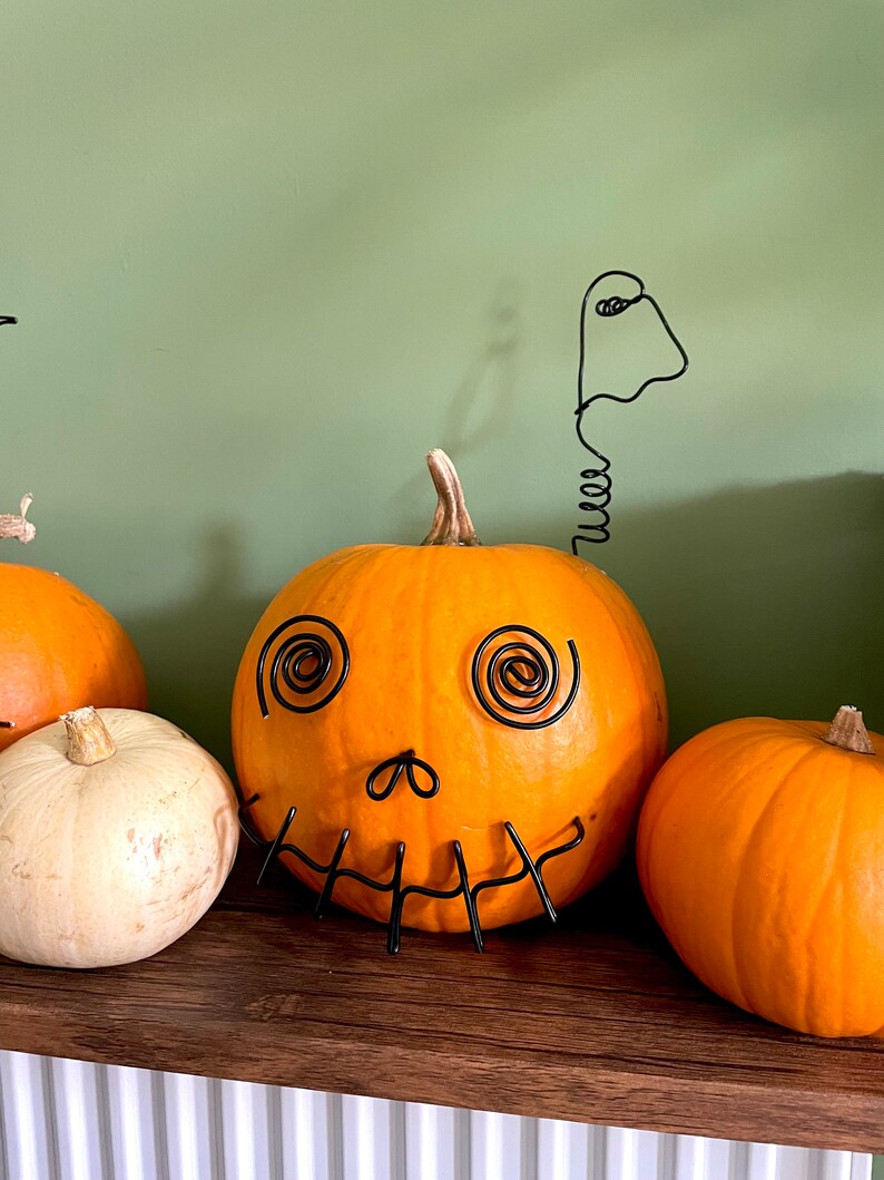 May include: A carved orange pumpkin with a wire ghost and a wire face on a wooden shelf. There are two other pumpkins on the shelf, one white and one orange.