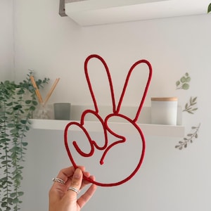 May include: A red rope sculpture shaped into a hand making a peace sign gesture. The sculpture is constructed from a single continuous length of thick red cord, forming the outline of the fingers and the circular base of the hand symbol.