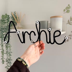 Puede incluir: Un letrero con el nombre "Archie" en cuerda negra y cursiva. El letrero tiene pequeños detalles de balones de fútbol. Una mano sostiene el letrero. El fondo incluye una estantería blanca con decoración y vegetación.