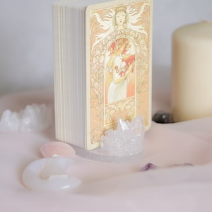 May include: A deck of tarot cards with a vintage illustration of a woman holding flowers. The cards are standing upright on a crystal stand with other crystals scattered around it.
