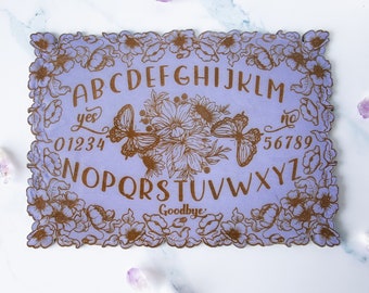 May include: A purple wooden Ouija board with a gold floral design and butterflies. The board features the alphabet, numbers 0-9, "yes", "no", and "goodbye".