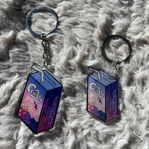 May include: Two keyrings with a juice box design. The boxes are blue and purple with a fruit illustration and the word "Fruity". Each keyring has a silver ring and chain.