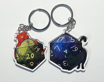 May include: Two keychains featuring a 20-sided die with a dragon and a cat. The dragon keychain has a red dragon with yellow eyes holding a green and black die with yellow numbers. The cat keychain has a black cat with white eyes holding a blue and purple die with white numbers.