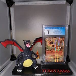 May include: A black and red Charizard Pokemon figure is displayed on a black stand with a CGC graded Charizard EX Pokemon card. The card is graded 8 and has a black star promo stamp. The stand has the text "TUBBYZARD" printed on it.