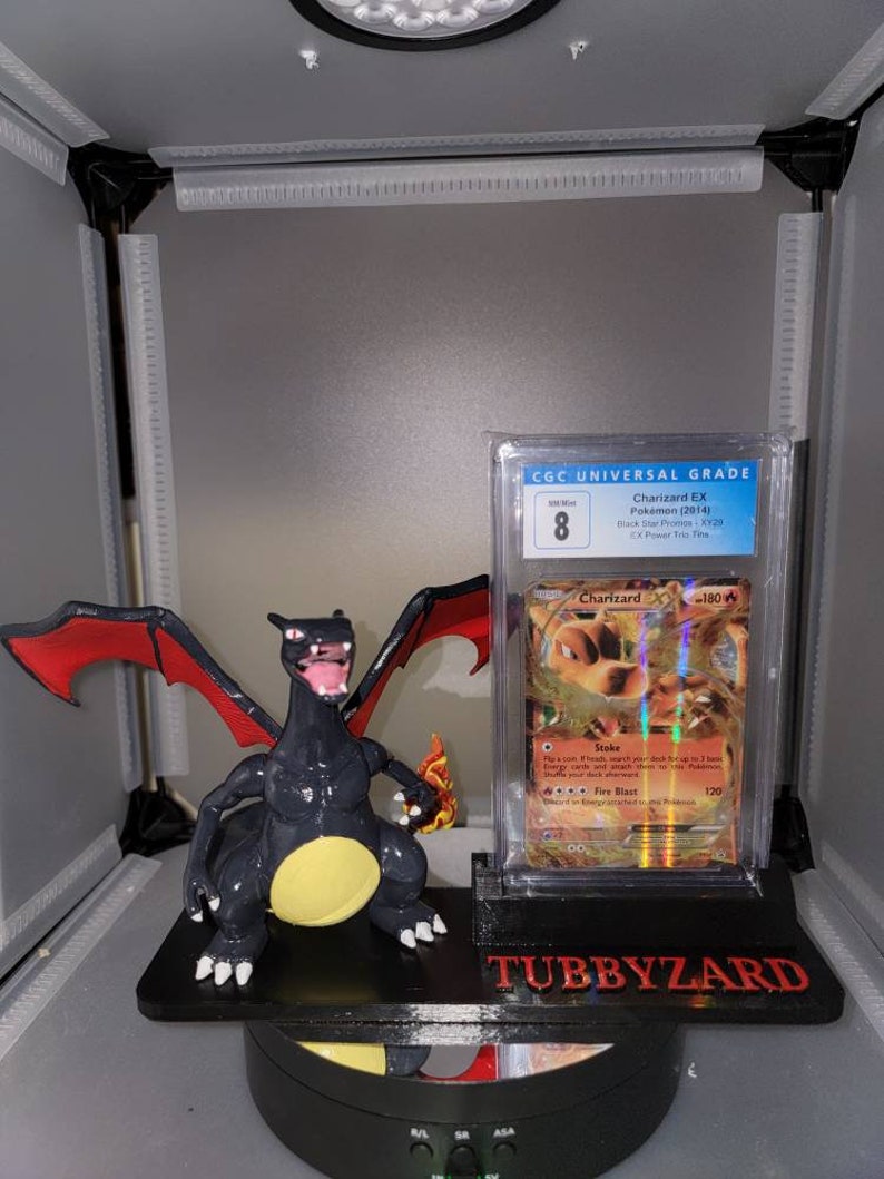 May include: A black and red Charizard Pokemon figure is displayed on a black stand with a CGC graded Charizard EX Pokemon card. The card is graded 8 and has a black star promo stamp. The stand has the text "TUBBYZARD" printed on it.