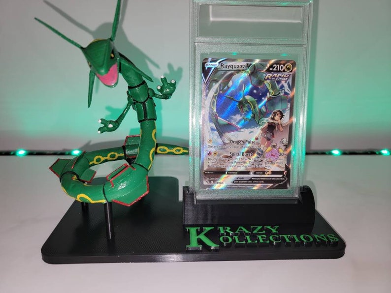 May include: A black plastic display stand with a green Rayquaza Pokemon figure and a graded Pokemon card featuring Rayquaza. The stand has the text "KRAZY KOLLECTIONS" printed on the front.