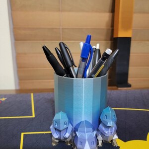 May include: A blue and silver 3D printed pen holder shaped like a Pokemon character. The holder is filled with pens and pencils.