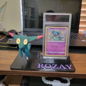 May include: A black card holder with a clear plastic case for a Pokemon card. The card is a Dreepy card with a blue and green creature on it. The card holder has the name "ROZAY" printed on it in purple letters. A green and red Pokemon figurine is in front of the card holder.