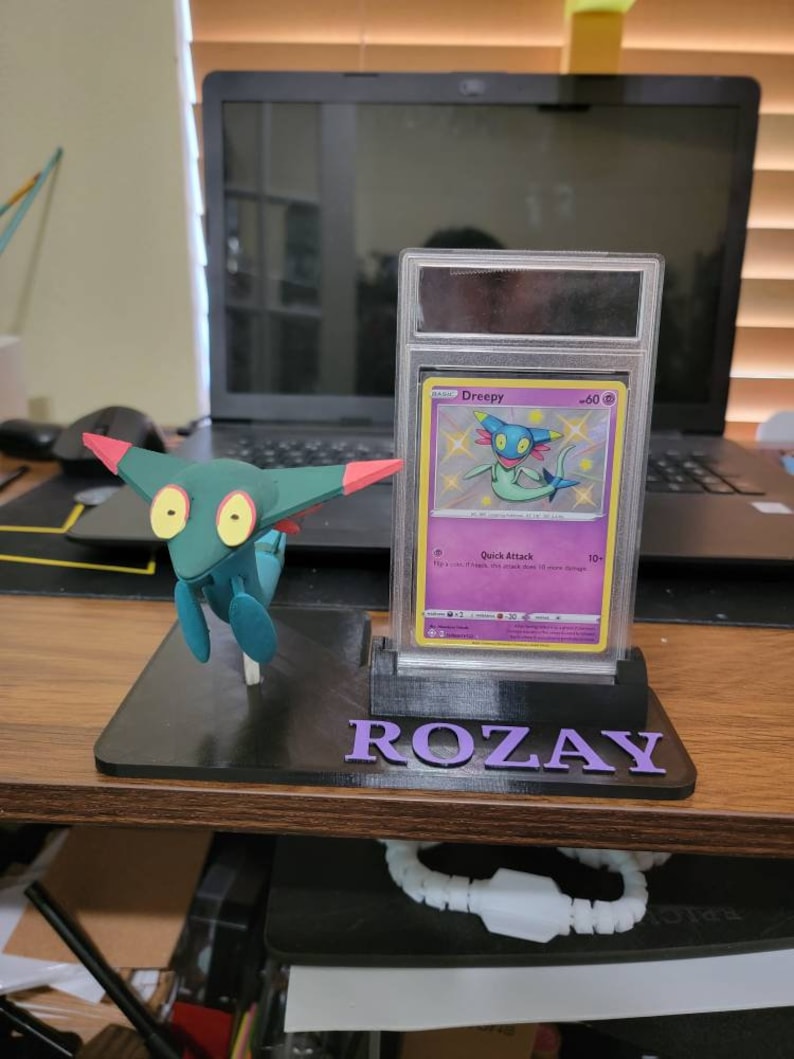 May include: A black card holder with a clear plastic case for a Pokemon card. The card is a Dreepy card with a blue and green creature on it. The card holder has the name "ROZAY" printed on it in purple letters. A green and red Pokemon figurine is in front of the card holder.