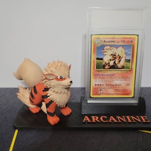 May include: A black plastic display stand with a clear plastic case holding a Pokemon trading card featuring Arcanine. The card is a holographic card with a red, orange, and yellow background. The card is titled "Arcanine" and has a picture of Arcanine on it. The stand has the word "Arcanine" printed on it in white letters. A small orange and brown Arcanine figurine is sitting on the stand.