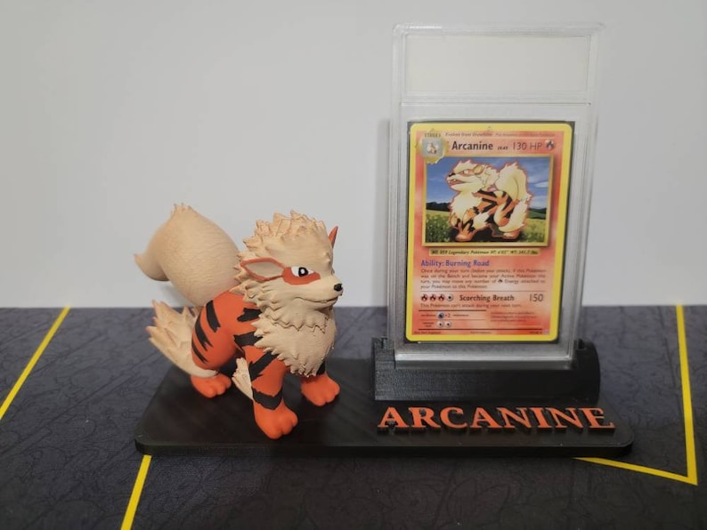 May include: A black plastic display stand with a clear plastic case holding a Pokemon trading card featuring Arcanine. The card is a holographic card with a red, orange, and yellow background. The card is titled "Arcanine" and has a picture of Arcanine on it. The stand has the word "Arcanine" printed on it in white letters. A small orange and brown Arcanine figurine is sitting on the stand.