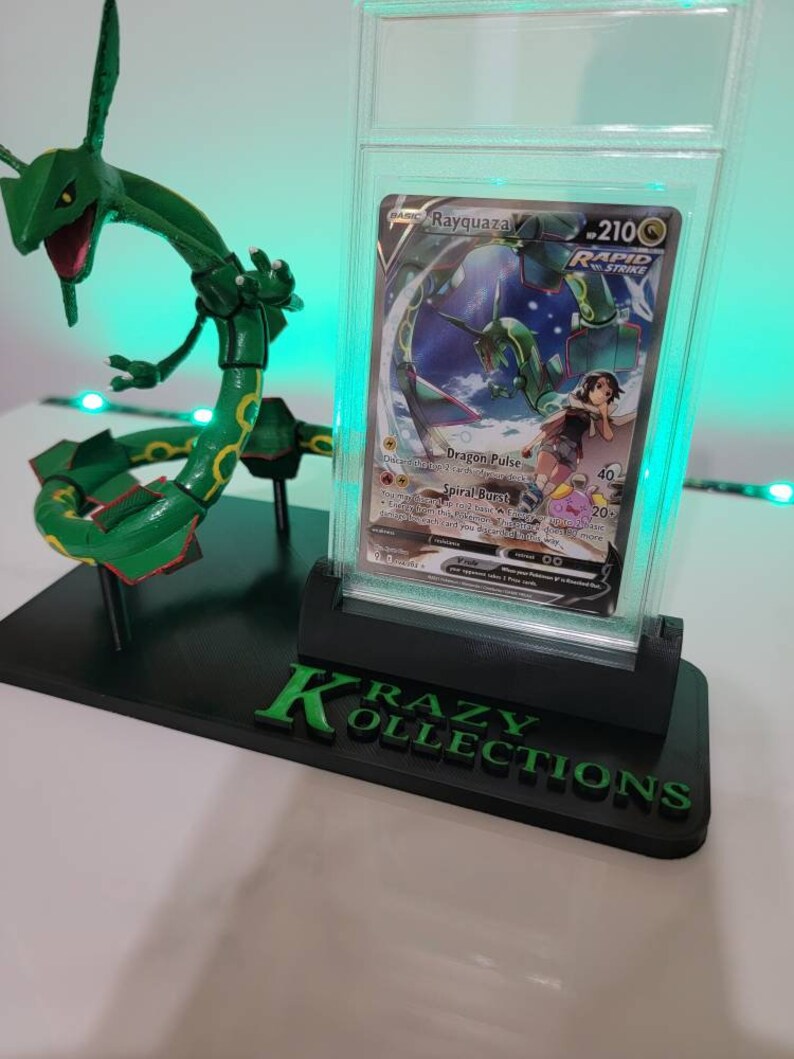 May include: A green and white Pokemon card featuring Rayquaza, a dragon-like Pokemon, in a clear plastic protective case. The card is on a black display stand with the words "Krazy Kollections" in green letters.
