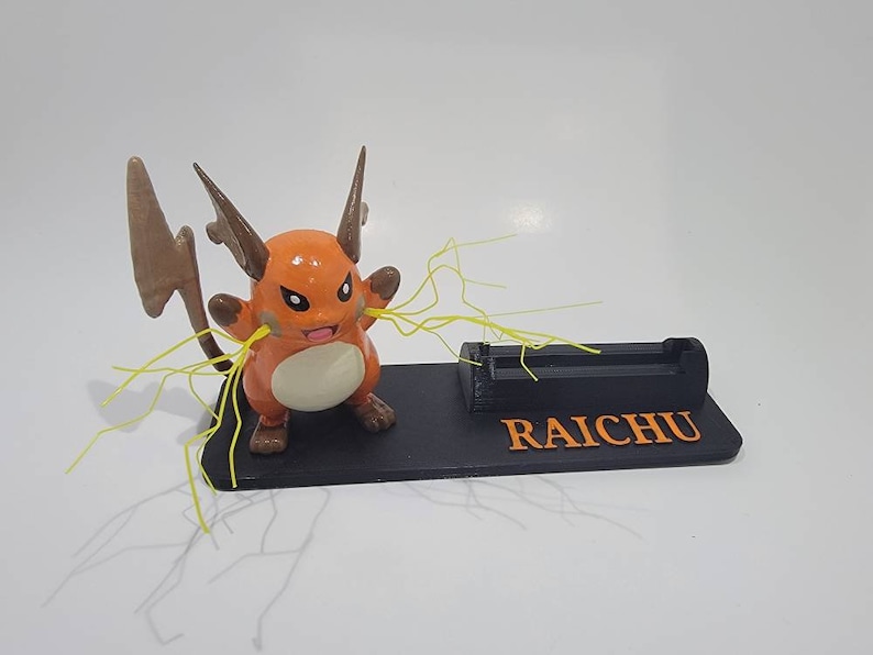 May include: A 3D printed phone stand featuring a cartoon Raichu Pokemon with yellow lightning bolts. The stand is black with the word "RAICHU" printed in orange.