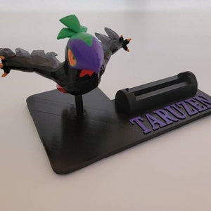 May include: A black and green 3D printed phone stand with a purple bird figurine on top. The stand has a slot for a phone and the word "TARUZEN" printed on the base.
