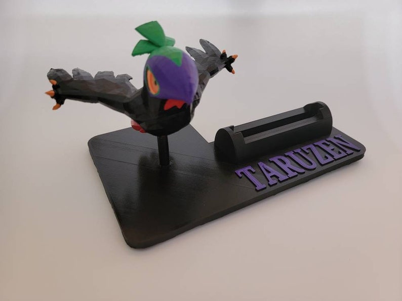 May include: A black and green 3D printed phone stand with a purple bird figurine on top. The stand has a slot for a phone and the word "TARUZEN" printed on the base.