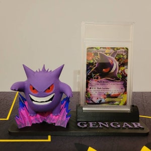May include: A 3D printed Gengar Pokemon figure stands on a black base with the word "Gengar" printed on it. A Gengar Pokemon card is displayed in a clear plastic case behind the figure.