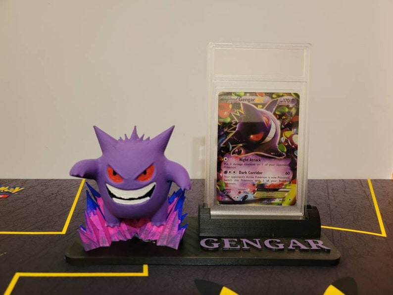 May include: A 3D printed Gengar Pokemon figure stands on a black base with the word "Gengar" printed on it. A Gengar Pokemon card is displayed in a clear plastic case behind the figure.
