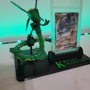 May include: A green and yellow articulated Pokemon figure of Rayquaza, posed in a circle, on a black display stand with a clear plastic case holding a Rayquaza Pokemon card. The card is graded with a 210 score and the stand has the text "KRAZY KOLLECTIONS" printed on it.