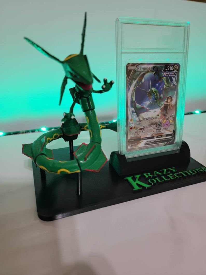 May include: A green and yellow articulated Pokemon figure of Rayquaza, posed in a circle, on a black display stand with a clear plastic case holding a Rayquaza Pokemon card. The card is graded with a 210 score and the stand has the text "KRAZY KOLLECTIONS" printed on it.