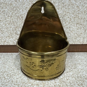 May include: A vintage brass wall-mounted container with a curved top and a circular base. The container features floral and leaf engravings around the base. It has a hole at the top for hanging.