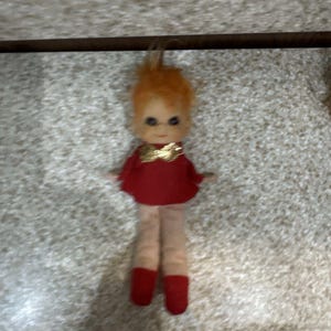 Puede incluir: Una muñeca pequeña con cabello naranja, ojos grandes y oscuros, y tono de piel claro. La muñeca viste una parte superior roja con un lazo dorado brillante en el cuello y botas rojas a juego. La figura tiene una expresión facial sencilla.
