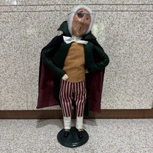 May include: A decorative figurine of a man with an eye patch, wearing a green coat with a burgundy lining, a gold shirt, and red and white striped pants. The figurine has a white wig and a white bow tie. It stands on a green base.