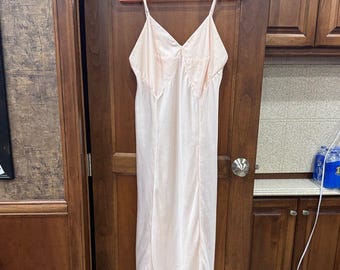 May include: A pale pink slip dress with spaghetti straps hangs on a pink hanger. The dress has a V-neck and a flowing, knee-length design. The fabric appears to be a soft, silky material.