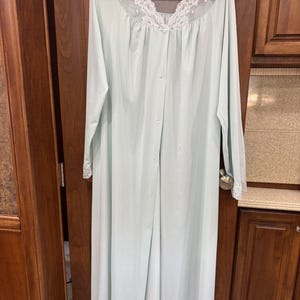 May include: A light blue, long-sleeved robe with a button-down front and a delicate lace collar and cuffs. The robe is hanging on a black hanger against a wooden door and cabinet background.