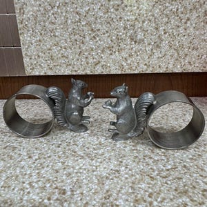 Könnte beinhalten: Zwei silberfarbene Serviettenringe mit Eichhörnchenfiguren. Jeder Ring hat ein kreisförmiges Band und eine detaillierte Eichhörnchenfigur. Die Eichhörnchen blicken sich an und halten etwas in ihren Pfoten. Der Hintergrund ist eine gesprenkelte Oberfläche.