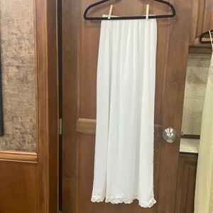 May include: A long, white slip dress hanging on a black hanger. The slip has a delicate lace trim at the hem. The garment is made of a lightweight, flowing fabric.