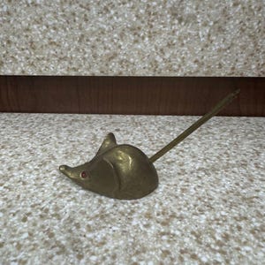Peut inclure: Une figurine de souris en laiton dotée d'une longue queue fine dirigée vers le haut et de petites pierres rouges incrustées en guise d'yeux. Le corps métallique présente une finition lisse et polie avec des oreilles pointues et un museau.