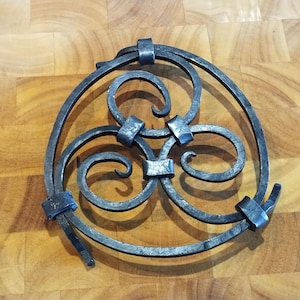 Op de afbeelding: Een handgemaakt decoratief stuk van smeedijzer met een cirkelvormig ontwerp. Het metaalwerk heeft drie wervelende scroll-elementen die zijn verbonden door vierkante accenten en een omlijstend cirkelvormig frame. Het stuk wordt op een houten oppervlak gepresenteerd.