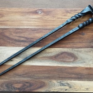 Könnte beinhalten: Zwei schwarze Metall-Kaminwerkzeuge mit gedrehten Griffen und hakenförmigen Enden, die auf einem Holzschneidebrett liegen. Die Werkzeuge sind angewinkelt, um ihr Design und ihre Handwerkskunst hervorzuheben. Die Holzmaserung des Bretts verleiht eine rustikale Note.