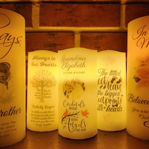May include: Five white pillar candles with different designs and text. The candles have a warm glow and are arranged in a row on a brick surface. The text on the candles includes "Always in Our Hearts", "Grandma Elizabeth", "The littlest feet leave the biggest footprints on our hearts", "Beloved Mom", and "Always Rever".