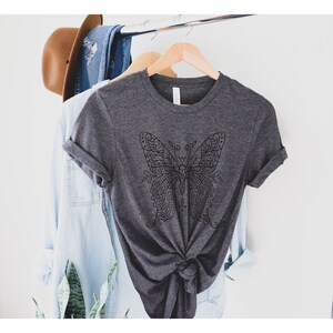 May include: A dark gray t-shirt with a black and white mandala-style butterfly design. The shirt is hanging on a hanger and is knotted at the bottom.
