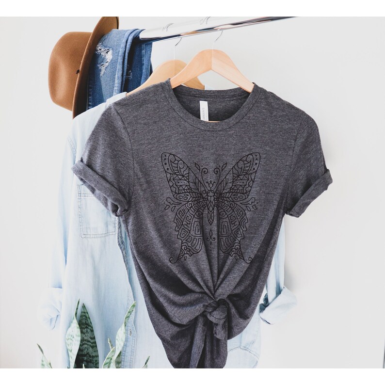 May include: A dark gray t-shirt with a black and white mandala-style butterfly design. The shirt is hanging on a hanger and is knotted at the bottom.