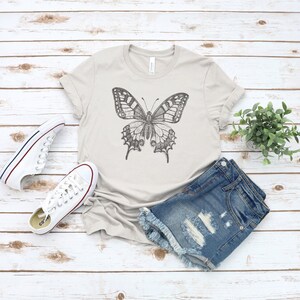May include: A light gray t-shirt with a black and white butterfly graphic. The t-shirt is paired with denim shorts and white sneakers.