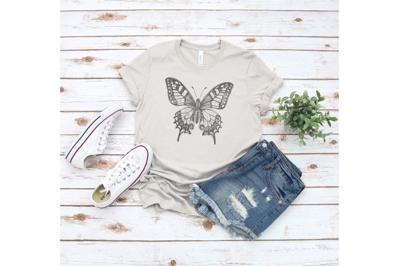 May include: A light gray t-shirt with a black and white butterfly graphic. The t-shirt is paired with denim shorts and white sneakers.