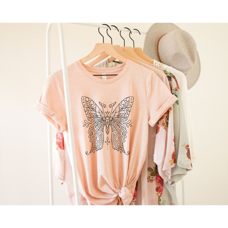 May include: A light pink t-shirt with a black and white mandala-style butterfly design. The shirt is on a hanger with other clothing.
