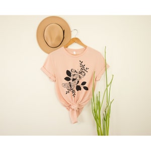 May include: A peach-colored t-shirt with a black and white graphic of two butterflies on a branch with leaves.