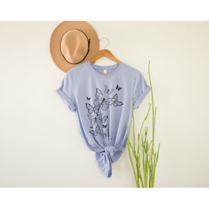 May include: A light blue t-shirt with a black butterfly graphic design. The butterflies are arranged in a cluster and appear to be flying upwards.