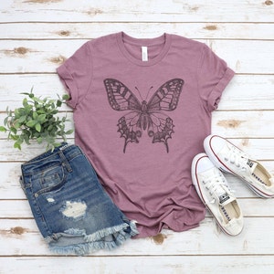 May include: A mauve t-shirt with a black and white butterfly graphic print. The shirt is laying flat on a white wood background with a pair of white canvas shoes and a pair of blue denim shorts.