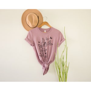 May include: A mauve t-shirt with a black butterfly graphic design. The shirt is tied at the front and is hanging on a wooden hanger.